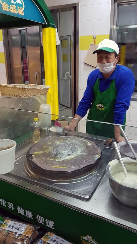 Vendor preparing jianbing on a round griddle behind glass