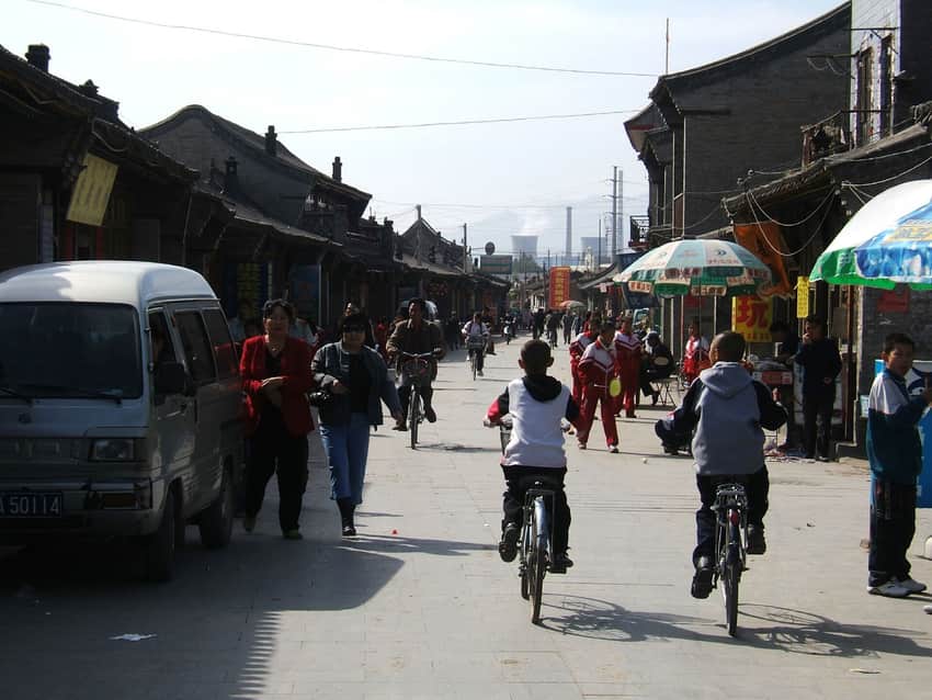 Dated busy old-city street with low buildings, bicycles, pedestrians and distant stacks