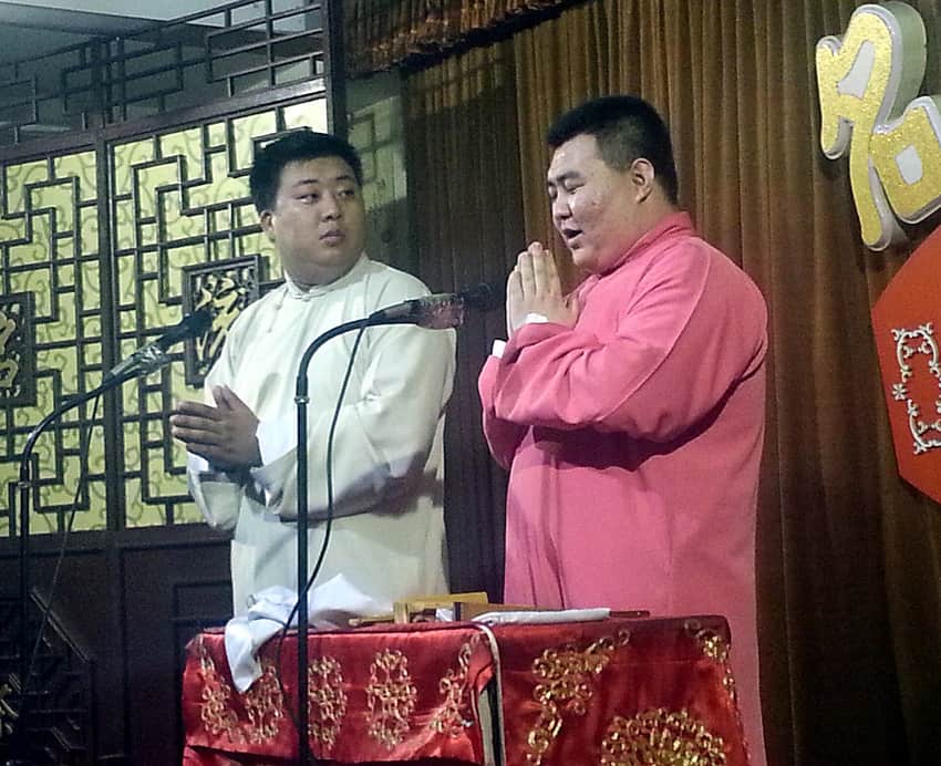 Two xiangsheng performers in long gowns behind a table and microphones in Tianjin