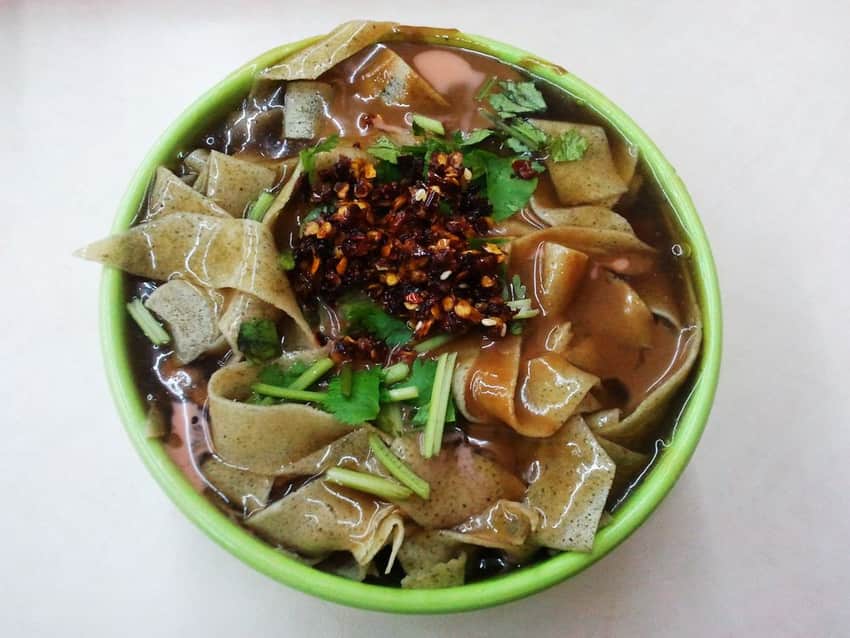 Top-down bowl of guobacai with cut crepe sheets, coriander and chili