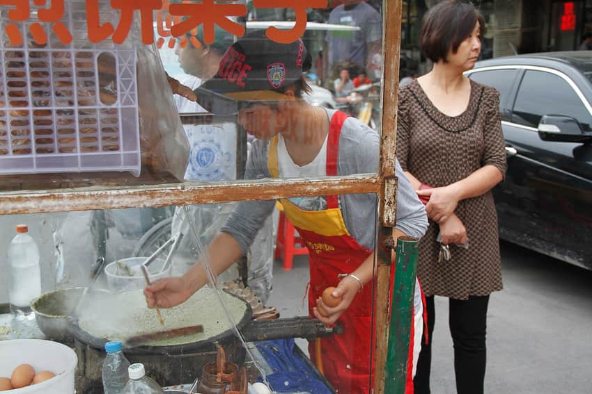 Street vendor spreading jianbing batter on a round griddle while holding an egg