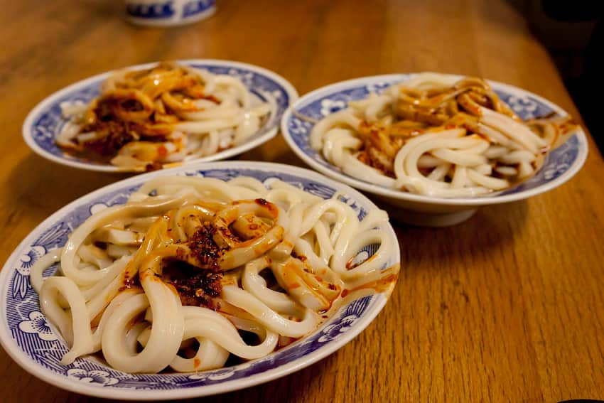 Three bowls of pale liangpi strips with dark sauce and chili oil