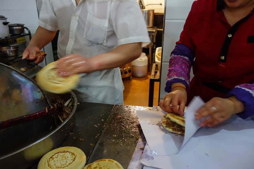 Two workers assembling roujiamo beside a heated pan